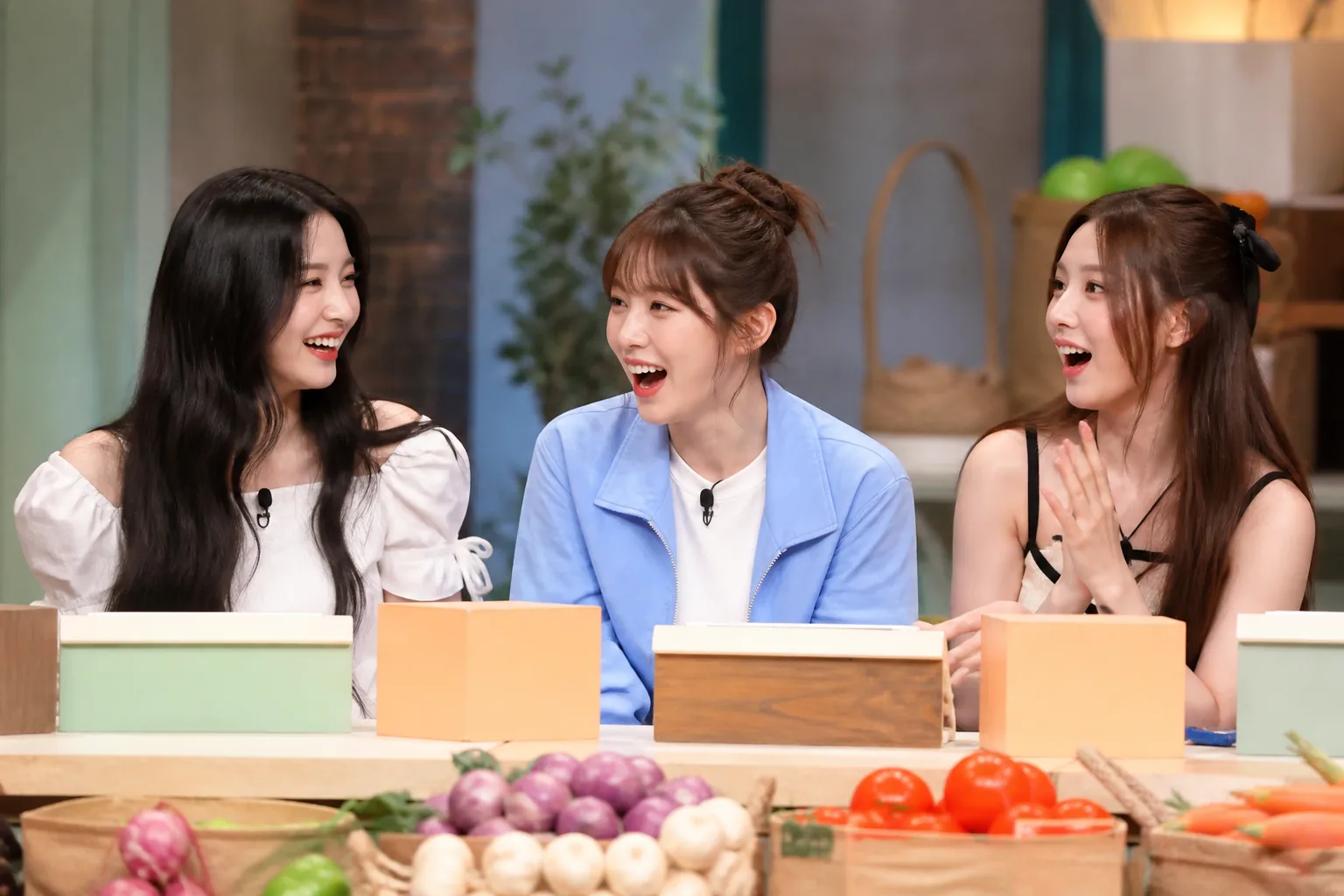 Young K-pop performers laughing together on a brightly lit variety-show set
